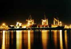 Containerschiff am Kai, 3 Kräne darüber, aufgenommen nachts vom Elbstrand aus
